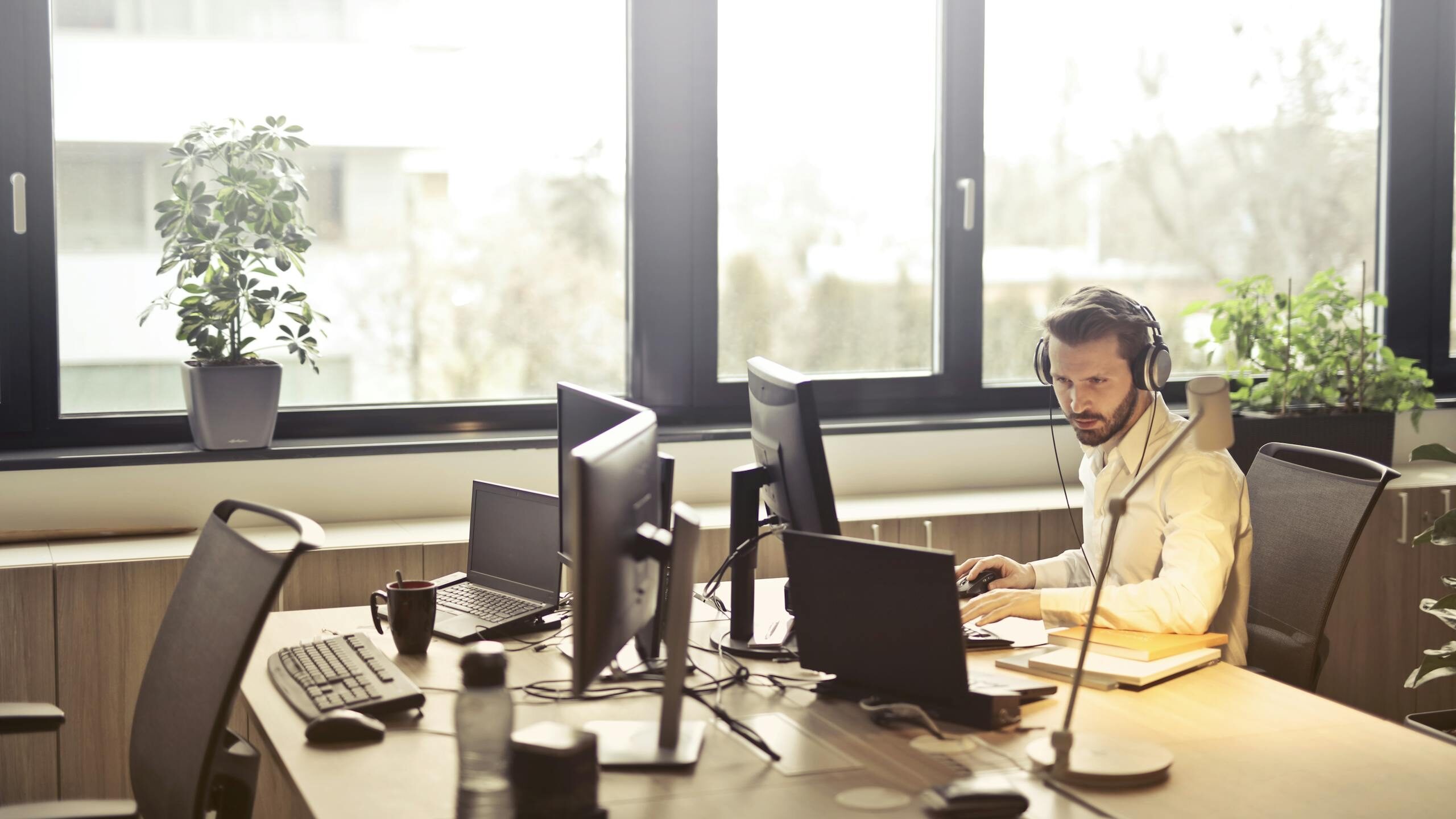 A businessman sits at a desk using multiple computers and a headset in a well-lit modern office.